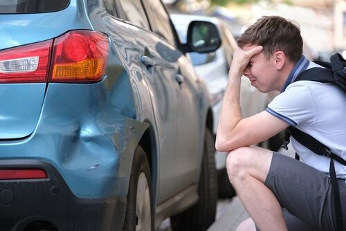 A distressed man crouching beside a damaged blue car, inspecting a dent on the rear corner panel, suggesting a hit-and-run accident.